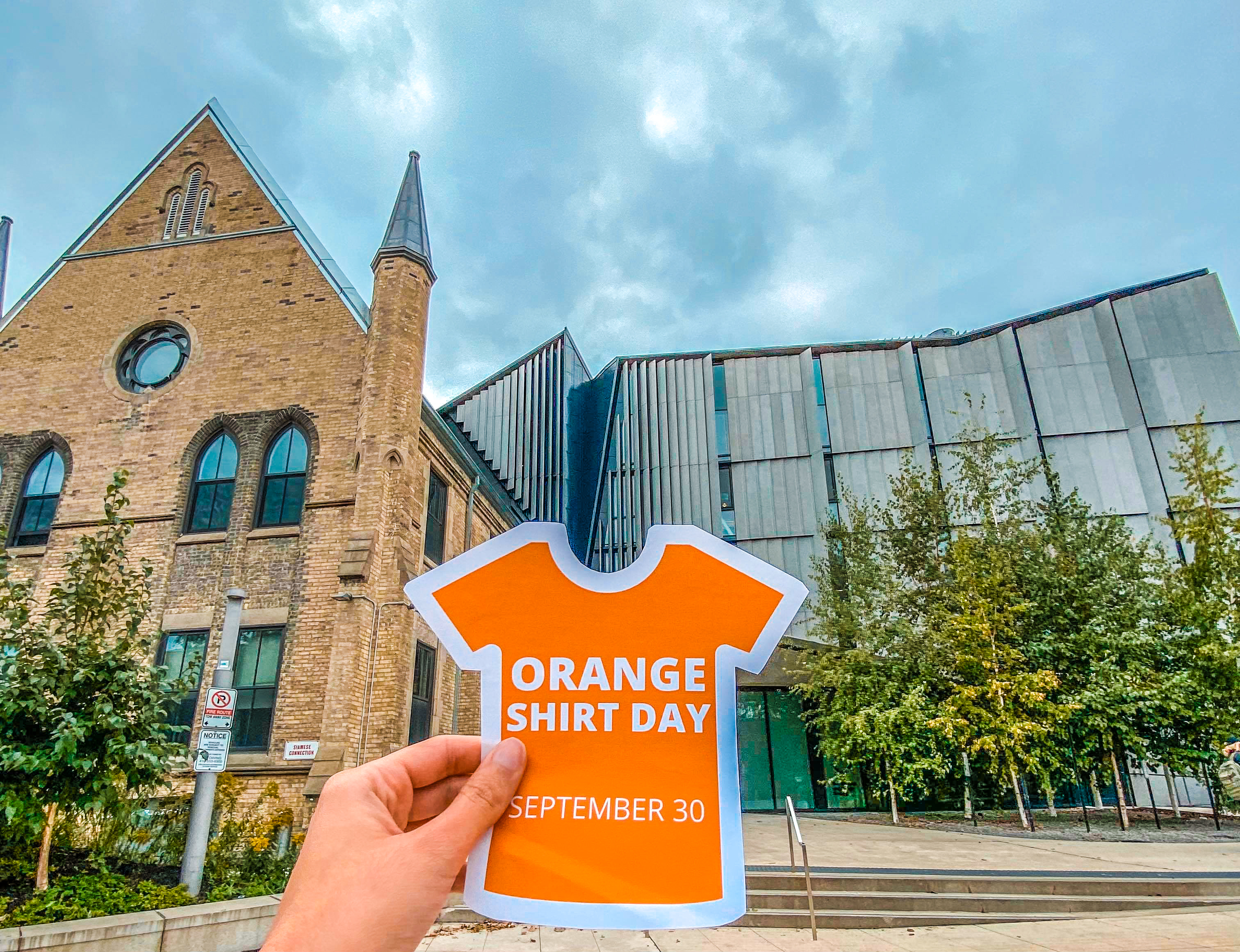 A hand holds an Orange Shirt Day sticker in front of One Spadina.
