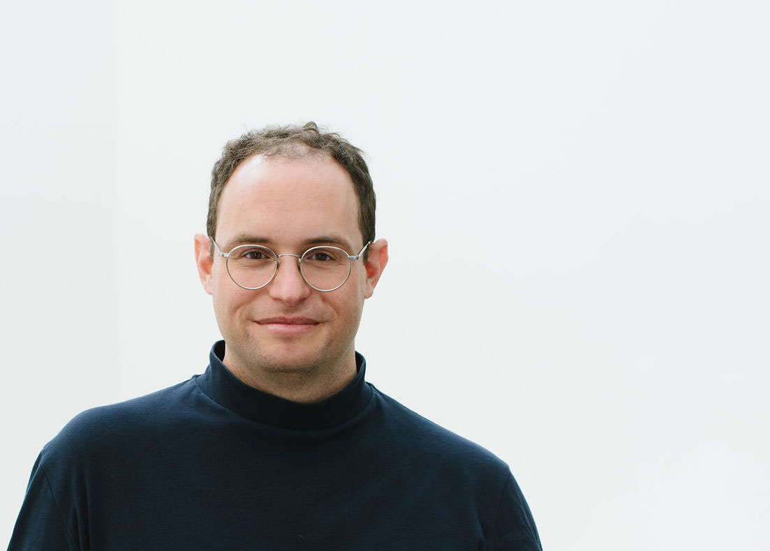 Portrait of Jon Cummings in a black mock neck sweater, from the chest up, standing in front of a gray background.